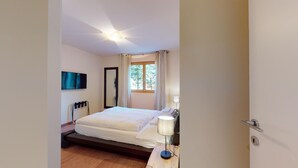 A bedroom with a bed, a bedside table with a lamp, a window with curtains, a mirror, and a painting on the wall. - Modern 3½ room flat on the ground floor, approx. (Madulain)