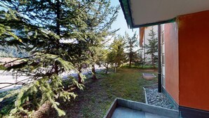 A modern building with a glass facade, a small outdoor seating area with a stone bench, and a well-maintained garden with various trees. - Modern 3½ room flat on the ground floor, approx. (Madulain)