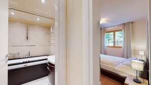 A hotel room with a bathroom featuring a bathtub, a sink, and a mirror. The room has a bed with a window, a bedside table, and a lamp. - Modern 3½ room flat on the ground floor, approx. (Madulain)