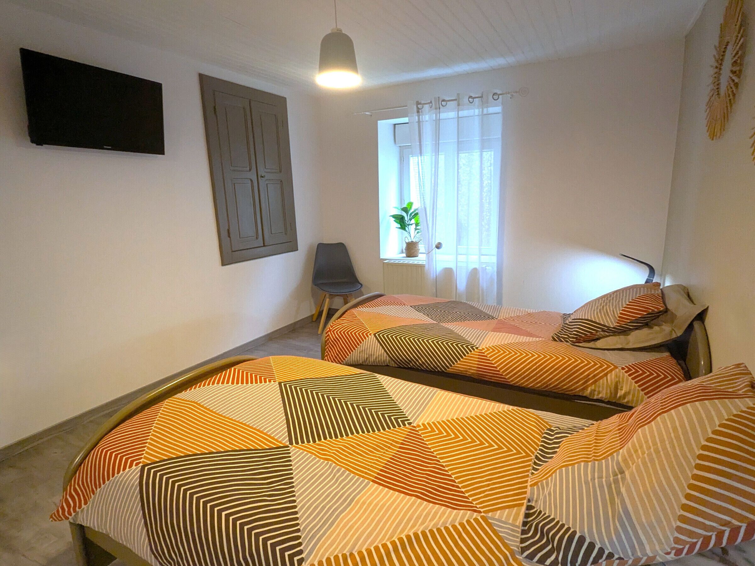 A bedroom with two beds, a window with curtains, a chair, and a TV mounted on the wall.