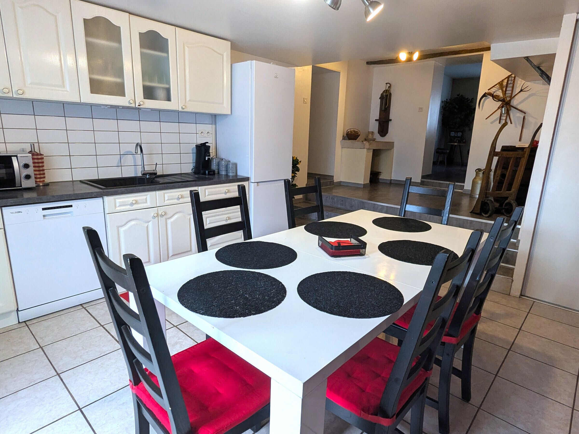 Cocina compacta con armarios blancos, lavavajillas y zona de comedor con mesa blanca y posavasos negros.