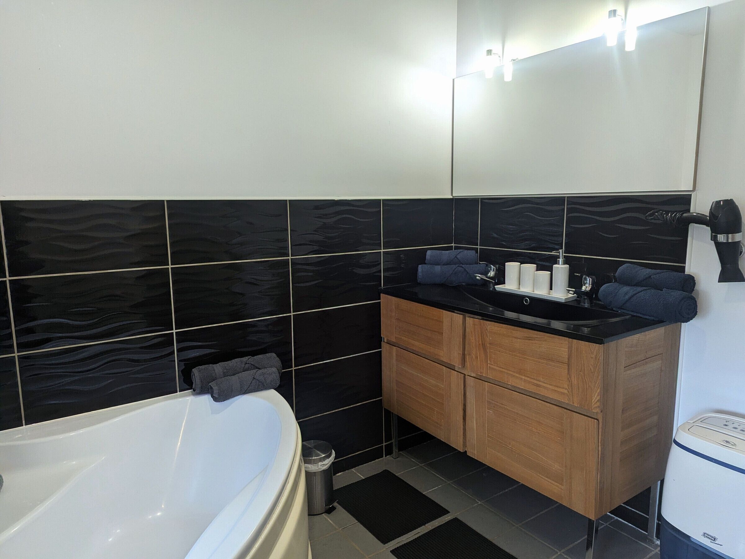 Une salle de bain moderne avec une baignoire blanche, des murs carrelés en noir et blanc, un meuble-lavabo en bois et un sèche-cheveux fixé au mur.