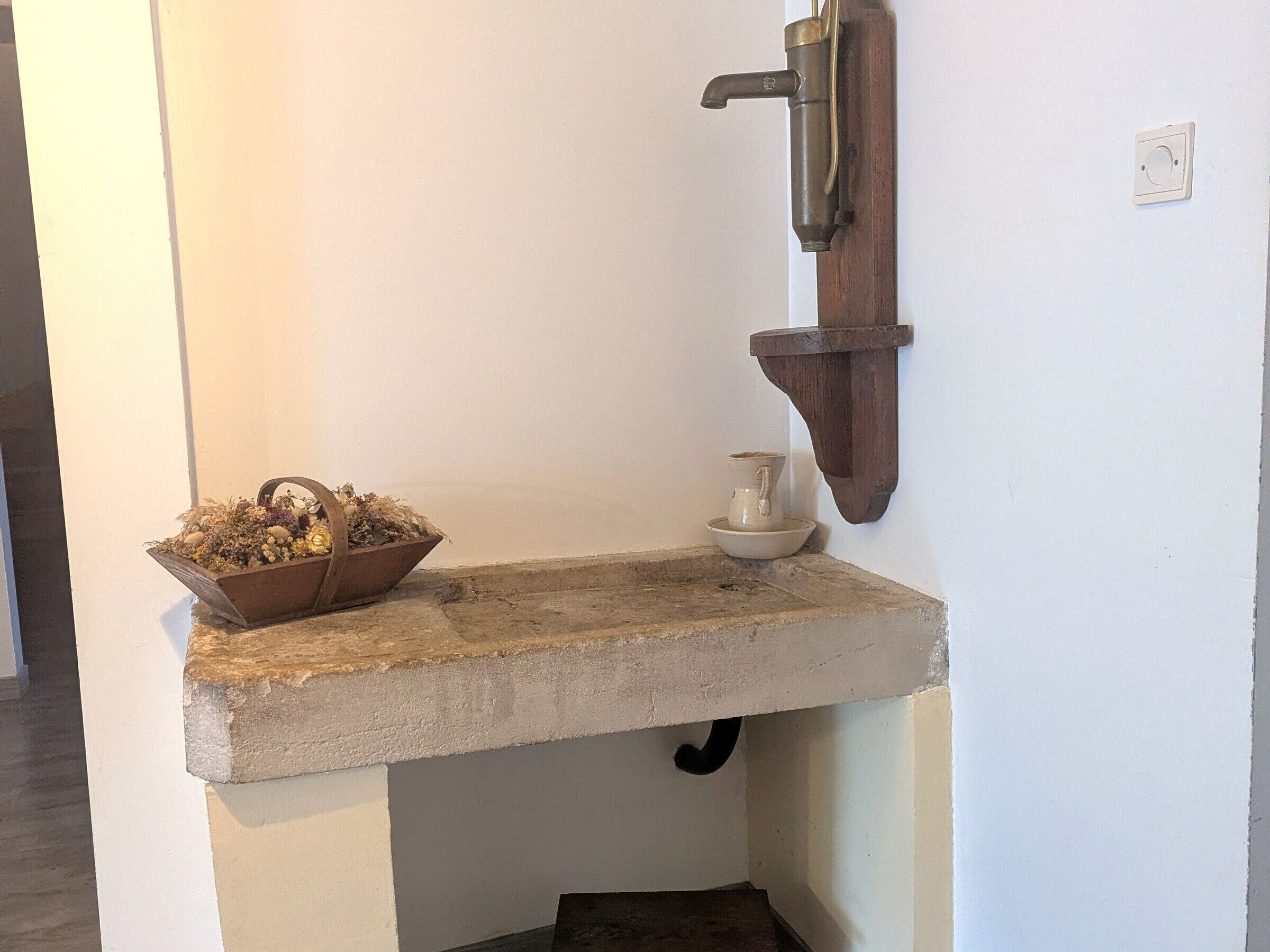 A rustic bathroom corner with a concrete sink, a wooden basin filled with flowers, and a small white pitcher on the counter.