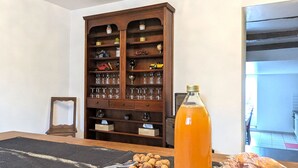 Un coin repas avec un buffet en bois rempli de verres et de petits objets décoratifs, une bouteille contenant un liquide orange posée sur la table, et un plateau de noix.
