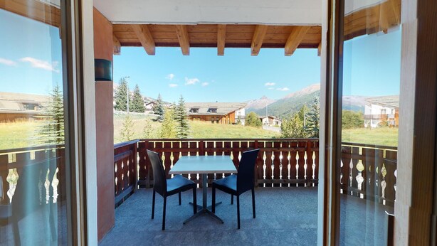 A balcony with a wooden table and chairs, overlooking a mountainous landscape with houses. - Modern 3½ room flat on the ground floor, approx. (Madulain)