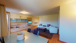 Una sala de estar moderna con un sofá blanco, una mesa de centro y un televisor. Incluye una zona de cocina con armarios y una mesa de comedor con sillas.
