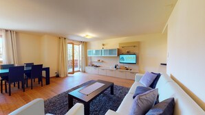 A modern living room with a sofa, coffee table, and dining area with chairs. There is a TV mounted on the wall and a balcony visible through the sliding doors. - Modern 3½ room flat on the ground floor, approx. (Madulain)