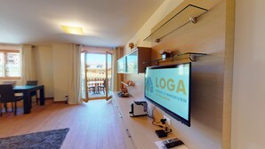 A modern living room with a flat-screen TV displaying 'LOGA Immobilien', a dining area with a table and chairs, and a view of a balcony with outdoor furniture. - Modern 3½ room flat on the ground floor, approx. (Madulain)