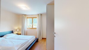 A hotel room with a bed, a bedside table with a lamp, a window with curtains, and a door. - Modern 3½ room flat on the ground floor, approx. (Madulain)