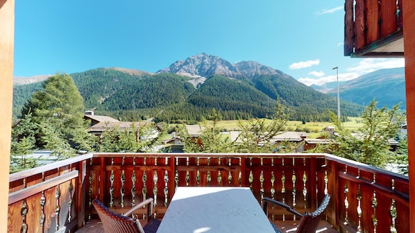 A balcony with a view of a mountain range, wooden railings, and a table with chairs. - Modern 3½ room flat on the ground floor, approx. (Madulain)