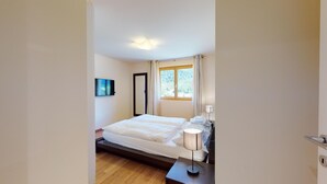 A bedroom with a bed, a bedside table with a lamp, a TV mounted on the wall, and a window with curtains. - Modern 3½ room flat on the ground floor, approx. (Madulain)