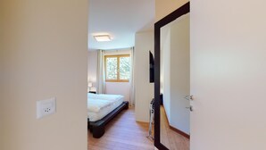A narrow hallway leading to a bedroom with a bed, a window, and a television mounted on the wall. - Modern 3½ room flat on the ground floor, approx. (Madulain)