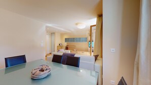 A modern hotel room with a glass dining table, a cozy seating area with a sofa and armchairs, and a kitchen area with cabinets and a countertop. - Modern 3½ room flat on the ground floor, approx. (Madulain)