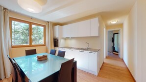 A compact kitchen with white cabinets, a central island with a sink, and a dining area with a glass table and chairs. - Modern 3½ room flat on the ground floor, approx. (Madulain)