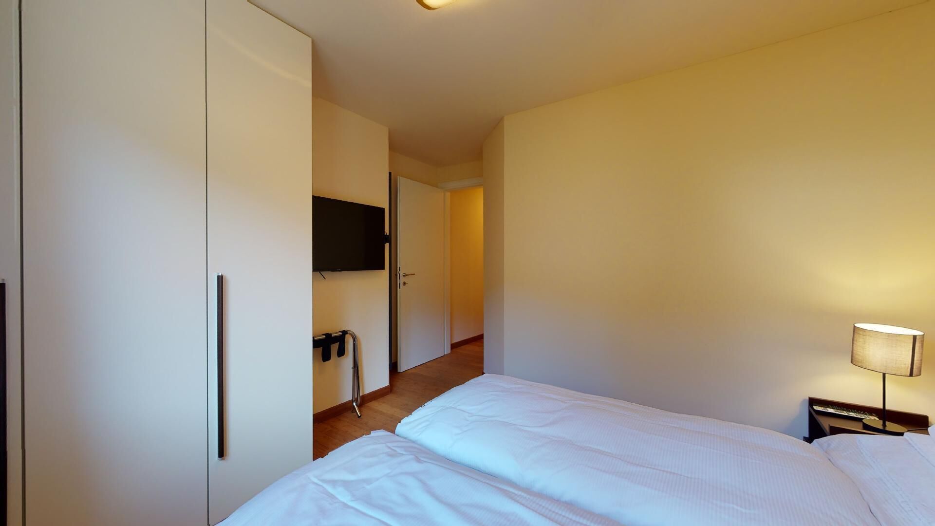 A hotel room with a bed, a TV mounted on the wall, a bedside table with a lamp, and a wardrobe.