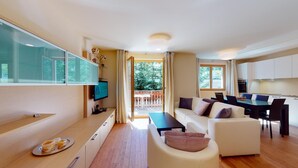 Een moderne woonkamer met een bank, een salontafel en een televisie. Er is een eethoek met een tafel en stoelen, en een keuken met kastjes en een raam.