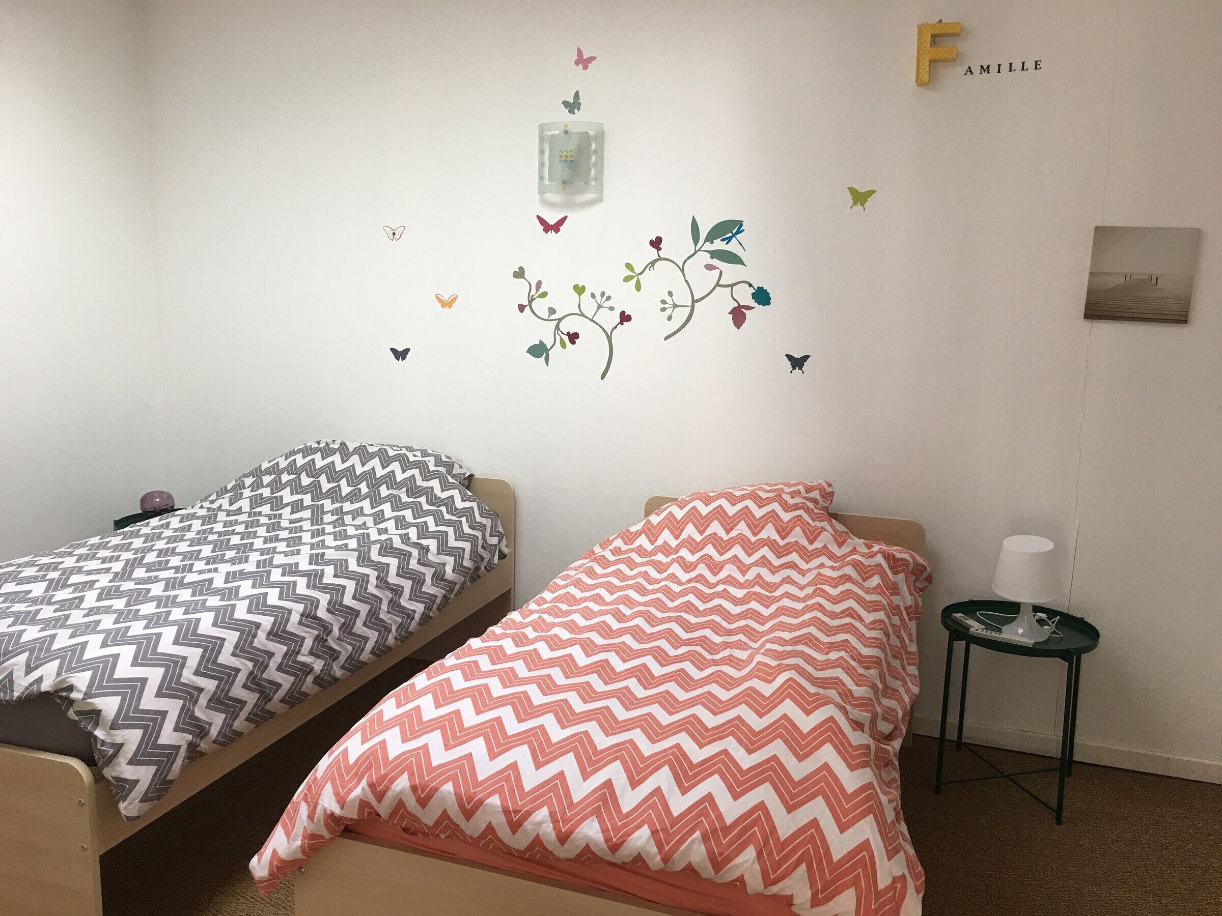A room with two beds, each with patterned bedding, a small table with a lamp, and a wall decal with butterflies and flowers.