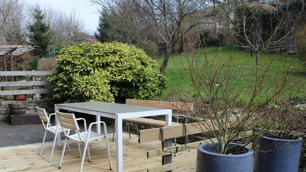 An outdoor wooden deck with a table and chairs, surrounded by greenery and trees.
