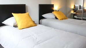 Ein Hotelzimmer mit zwei Betten, jeweils mit weißer Bettwäsche und einem gelben Kissen. An beiden Seiten der Betten befinden sich Nachttischlampen.