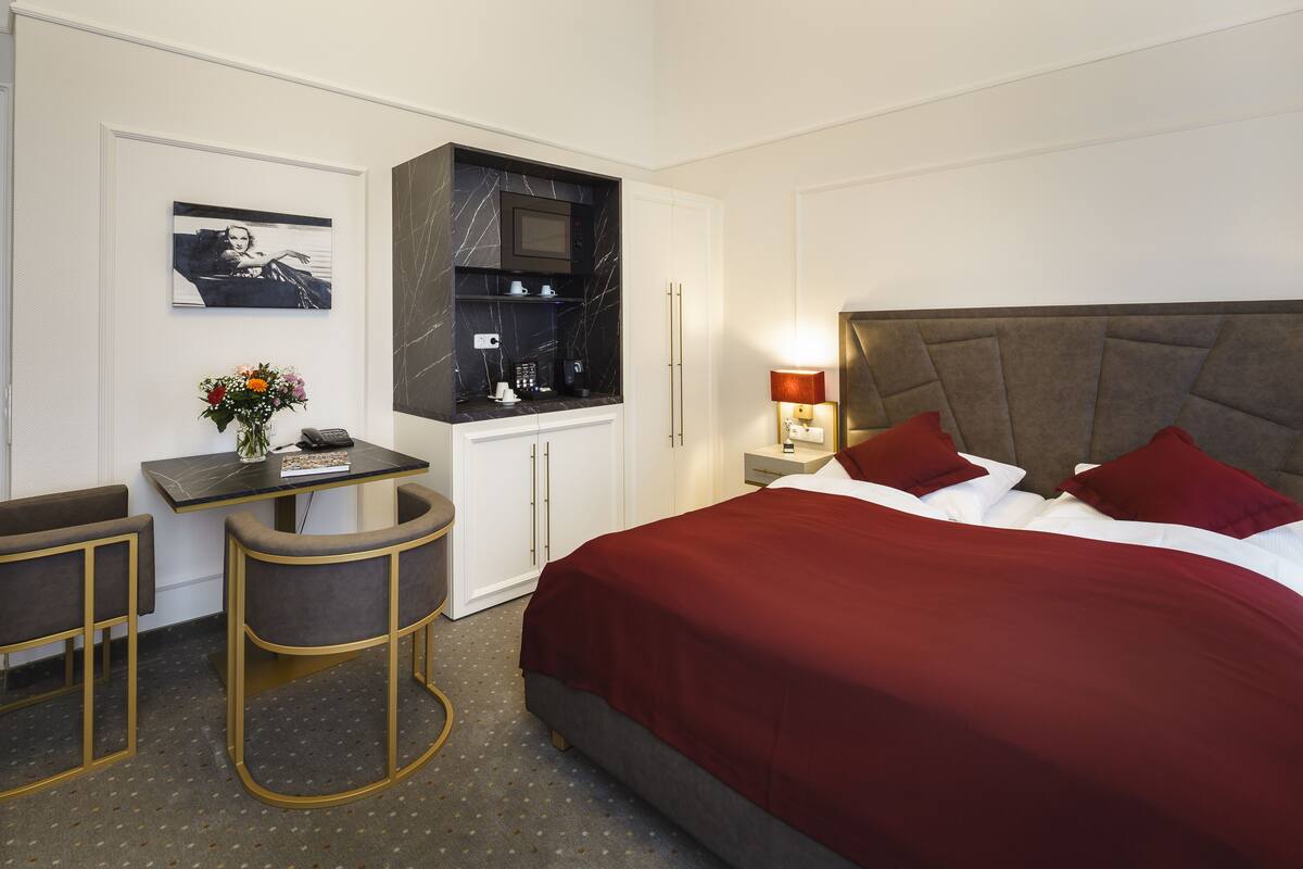 Une chambre d’hôtel comprenant un lit, un bureau avec une chaise, une petite table avec un vase de fleurs, un four à micro-ondes et une table de chevet avec une lampe.
