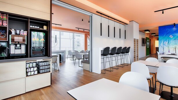 A modern dining area with white tables and chairs, a coffee station with dispensers, and a large window providing natural light. - B&B Hotel Rostock-Hafen (Rostock)