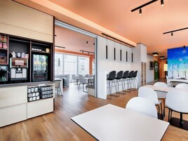 A modern dining area with white tables and chairs, a coffee station with dispensers, and a large window providing natural light.