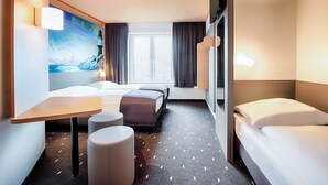 A hotel room with a bed, a desk, a chair, a small ottoman, a TV, and a window with curtains. - B&B Hotel Rostock-Hafen (Rostock)