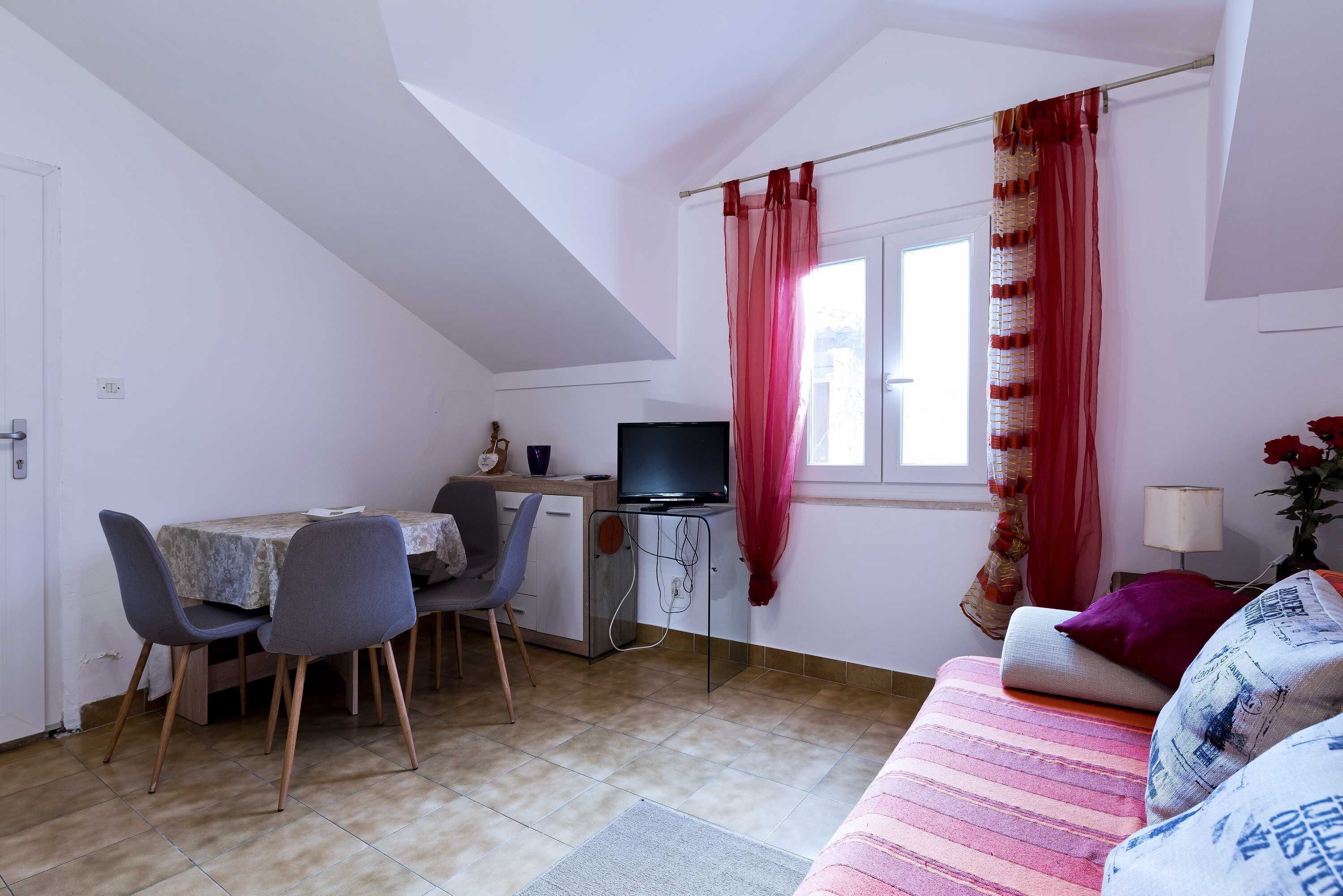 A compact room with a dining table, two chairs, a TV, a sofa, and a window with curtains.