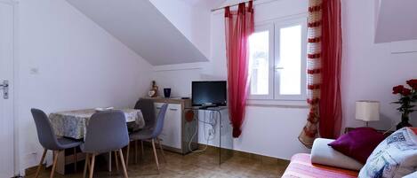A compact room with a dining table, two chairs, a TV, a sofa, and a window with curtains.