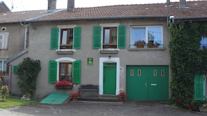 A two-story building with green shutters, a white door, and a green garage door. There are flower pots on the ground floor and a bench outside. - Charming Renovated Farmhouse, Terrace, Garden, Near Lakes & Nature, Close to Baccarat & Badonviller (Reherrey)