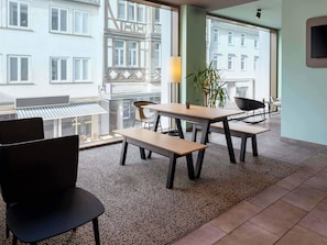 Un interior moderno con zona de comedor que incluye una mesa, sillas, una planta en maceta y vistas de un edificio a través de amplios ventanales.