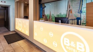A reception area with a B&B Hotels sign, a reception desk, a potted plant, and a view of the outside.