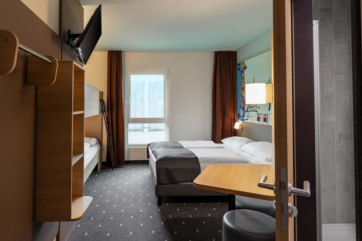 A hotel room with a bed, a desk, a chair, a TV mounted on the wall, and a window with curtains.