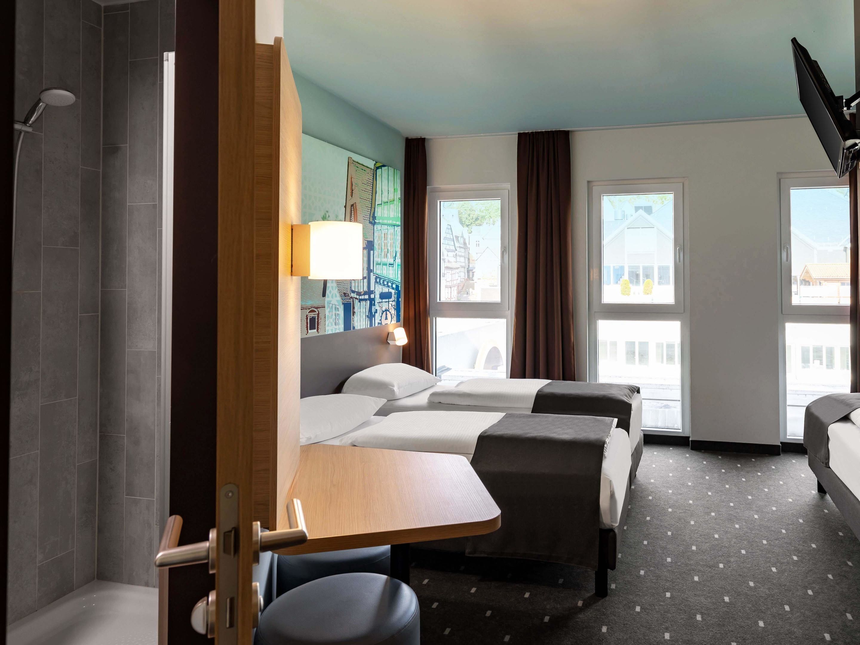 Een hotelkamer met twee bedden, een douche, een klein tafeltje en uitzicht op gebouwen door grote ramen.