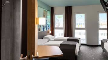 Een hotelkamer met twee bedden, een douche, een klein tafeltje en uitzicht op gebouwen door grote ramen.