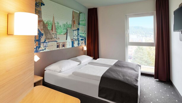 A hotel room with a bed, a desk, a painting on the wall, and a window with curtains.