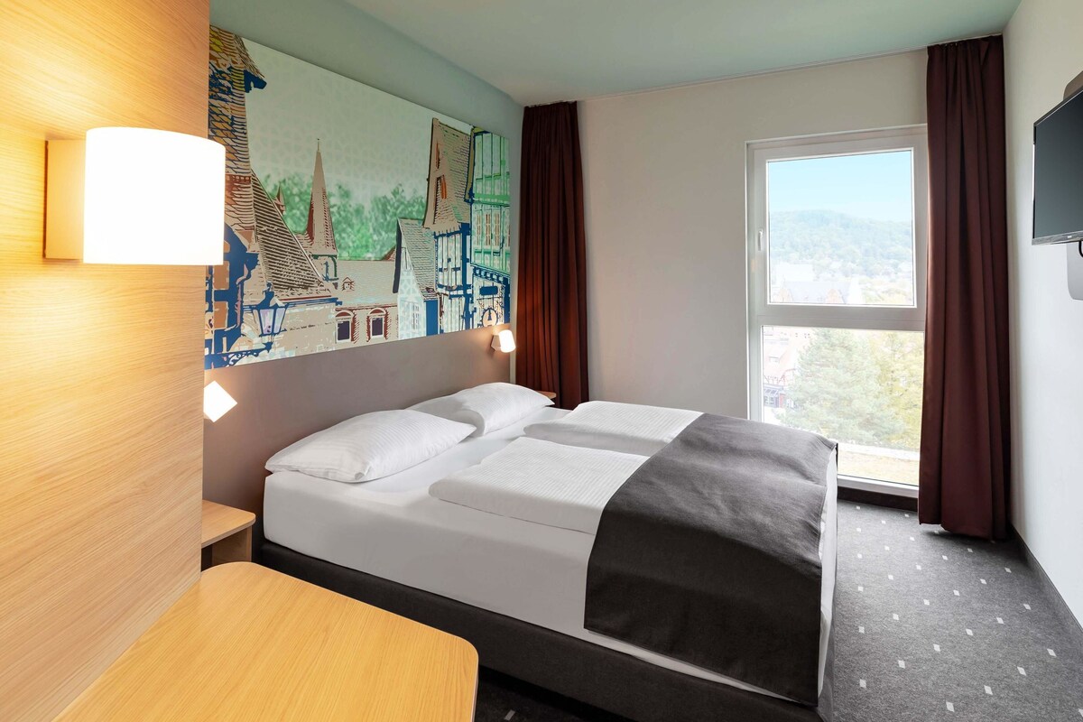 A hotel room with a bed, a desk, a painting on the wall, and a window with curtains.