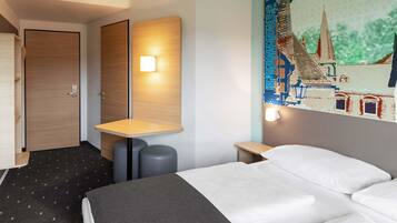 Habitación de hotel con cama, escritorio, silla, puerta y un cuadro de paisaje urbano en la pared.