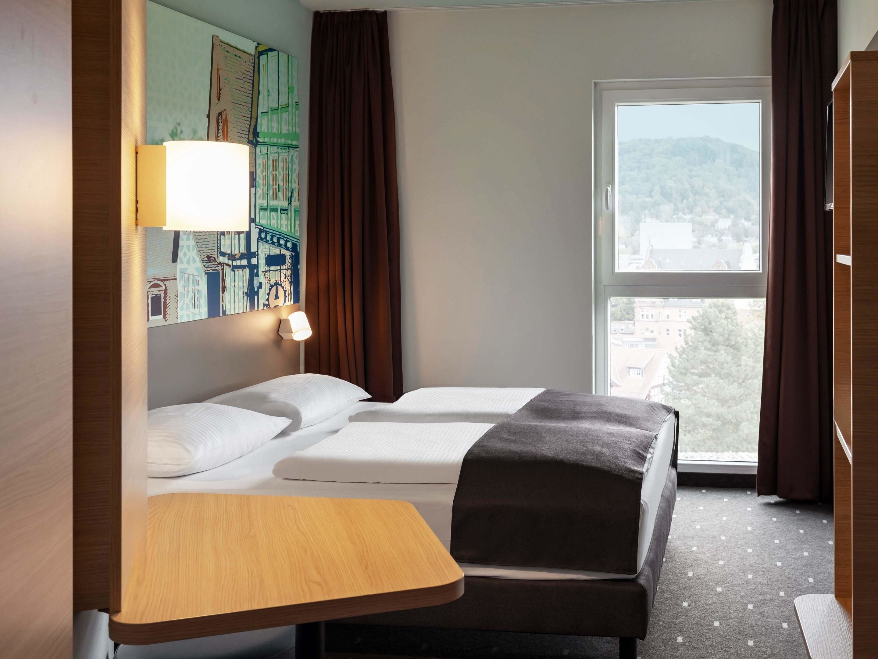 A hotel room with two beds, a wooden table, a lamp, and a large window with a view of buildings.