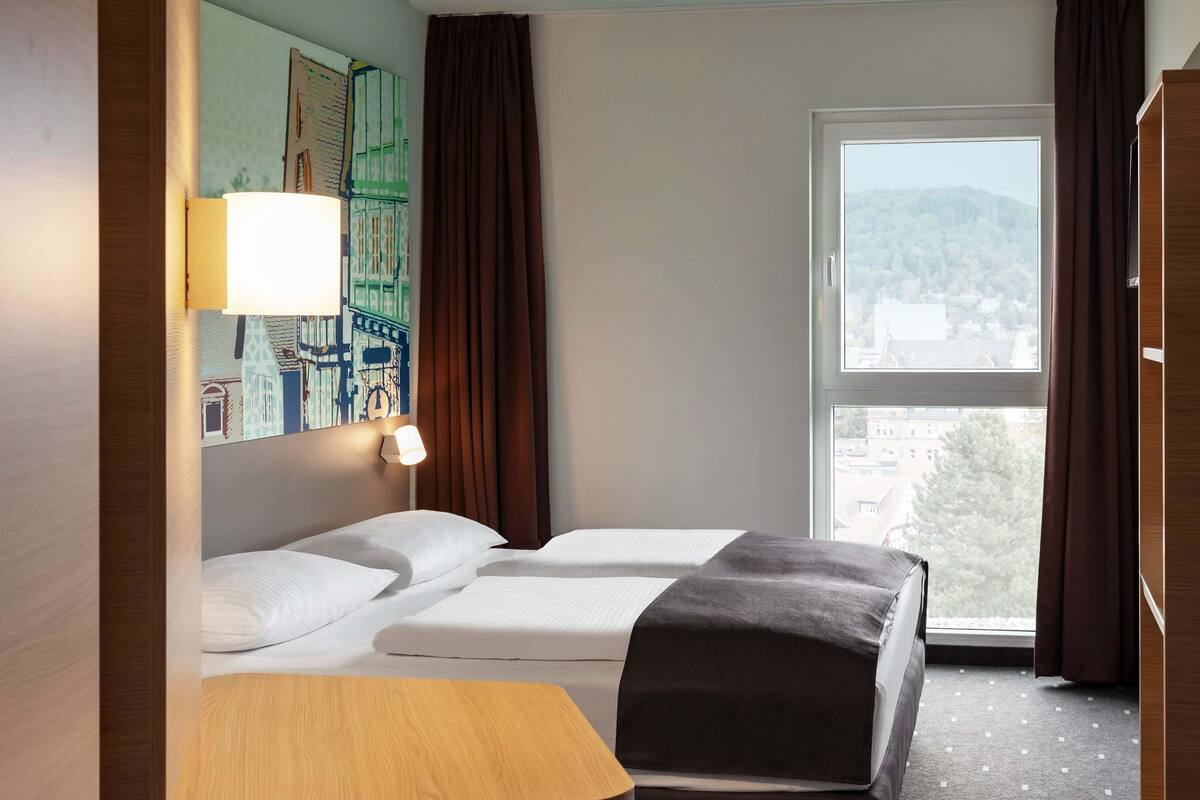 Habitación de hotel con dos camas, una mesa de madera, una lámpara y un ventanal con vistas a edificios.