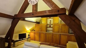 Une pièce mansardée chaleureuse avec un plafond en bois, un canapé, une télévision et un coin cuisine doté de meubles en bois et d’une lampe.