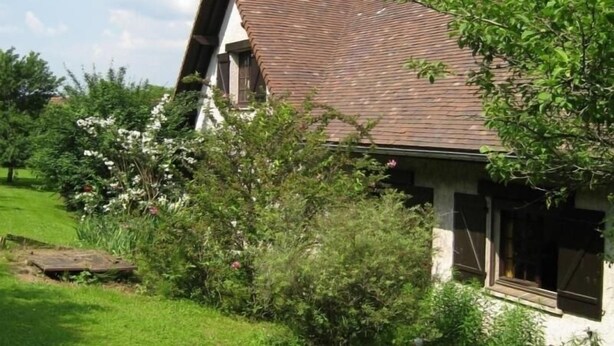 Une maison traditionnelle avec un toit en pente, entourée d’une végétation luxuriante et de plantes en fleurs.