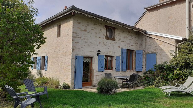 Una casa de piedra rústica con puerta de madera, contraventanas azules y un peque ño balcón. Hay sillas y una mesa en el jardín delantero.