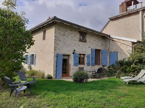 Une maison de campagne en pierre, avec une porte en bois, des volets bleus et un petit balcon. On y trouve des chaises et une table à l’extérieur, dans le jardin.