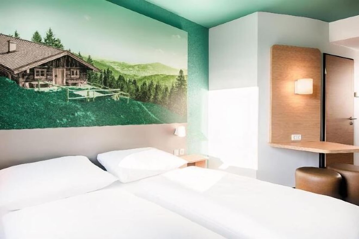 Habitación de hotel con cama, mesita de noche, lámpara de pared y un cuadro de una cabaña en la montaña.