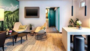 Una habitación de hotel moderna con una gran televisión de pantalla plana montada en la pared, un área de descanso con dos sillas y una mesita, y un comedor con una mesa blanca y taburetes negros.
