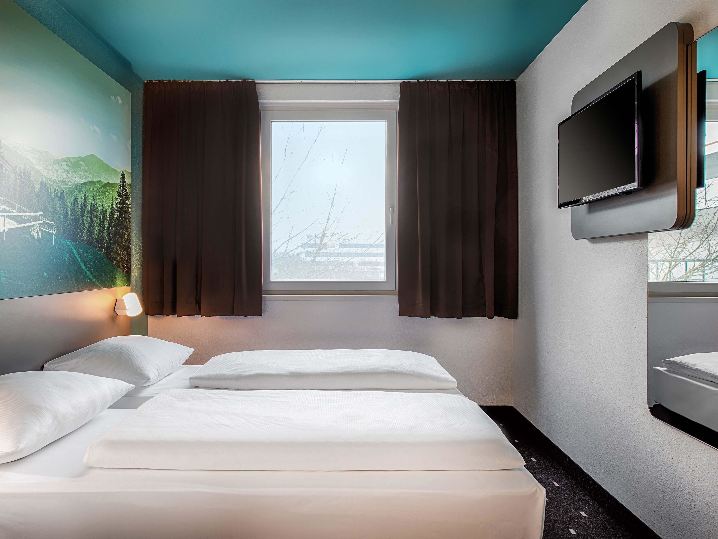 Une chambre d’hôtel avec un lit, un téléviseur fixé au mur, une fenêtre avec des rideaux et un tableau accroché au mur.