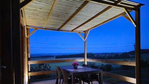 Un espace extérieur abrité, doté d’une table et de chaises en bois, offrant une vue sur la campagne et surmonté d’une structure de toit en métal.