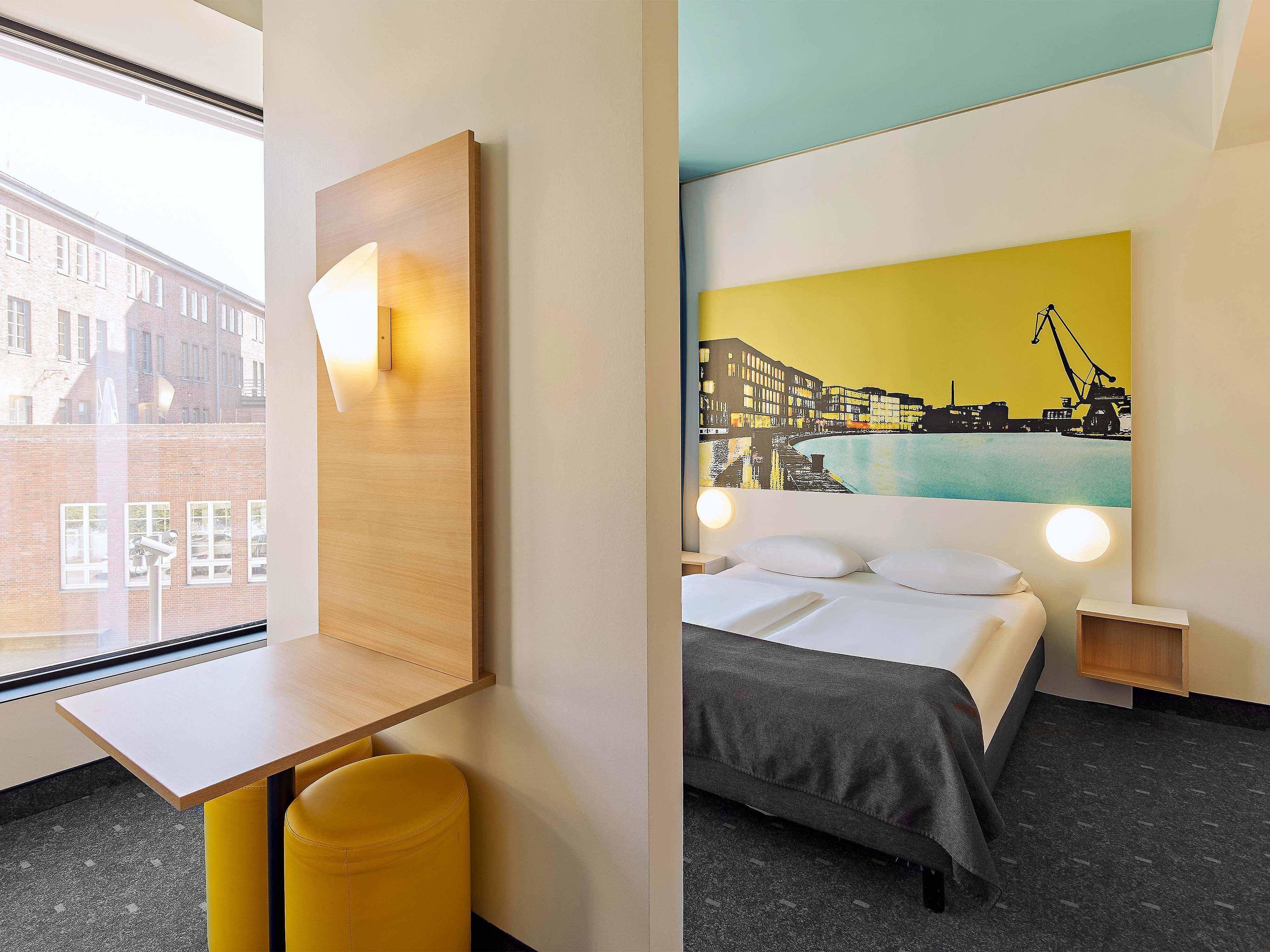 A hotel room with a bed, a desk, a chair, and a large window.