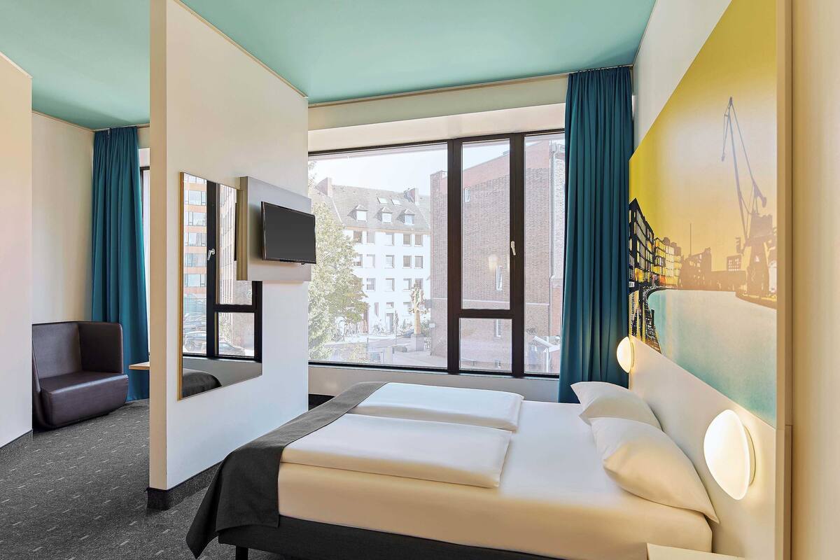 A hotel room with a bed, a TV, a desk, a chair, and large windows with a view of buildings.